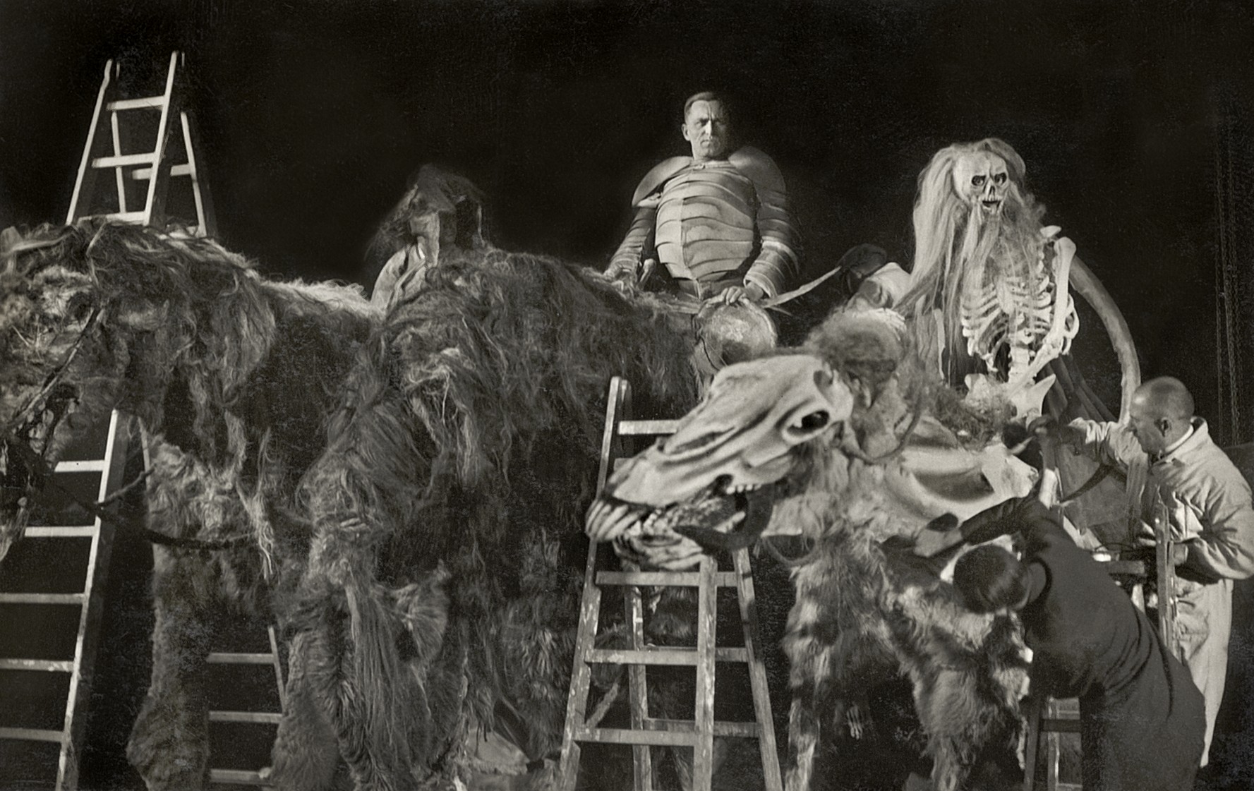 Crew members stand on stepladders around giant shaggy model horses on a dark film set, one ridden by a man in armour and another by a skeleton with long white hair, as technicians adjust the props.