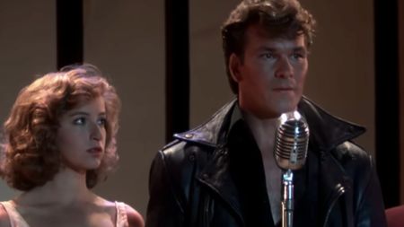 Jennifer Grey on the right in a pink dress, next to Patrick Swayze in leather and talking into a microphone in the last scene of Dirty Dancing
