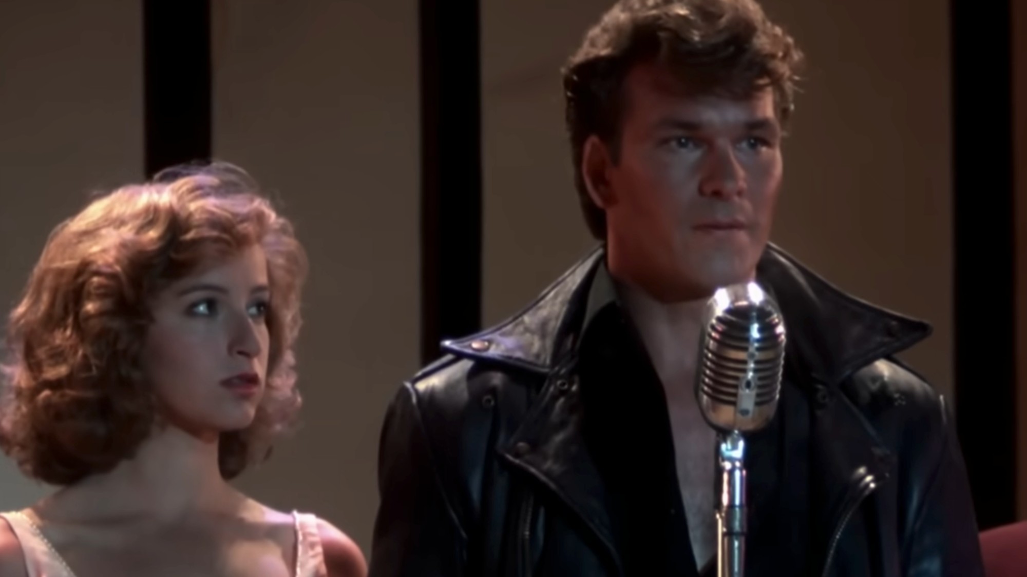 Jennifer Grey on the right in a pink dress, next to Patrick Swayze in leather and talking into a microphone in the last scene of Dirty Dancing