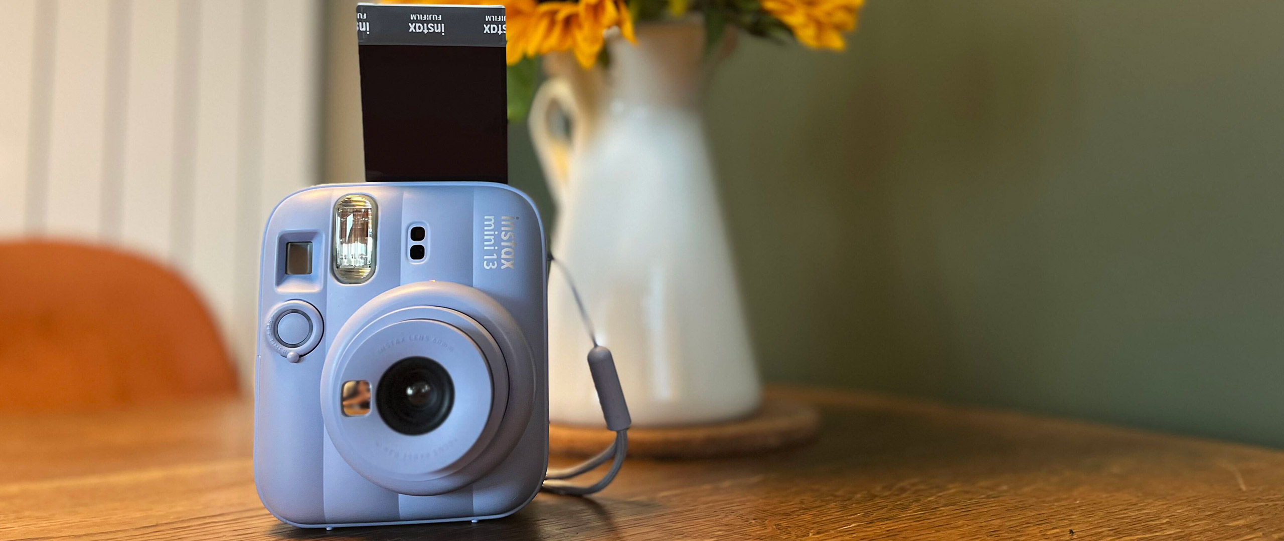 The Frost Blue Instax Mini 13 on a table with a print sticking out of the top of it next to a vase of sunflowers 