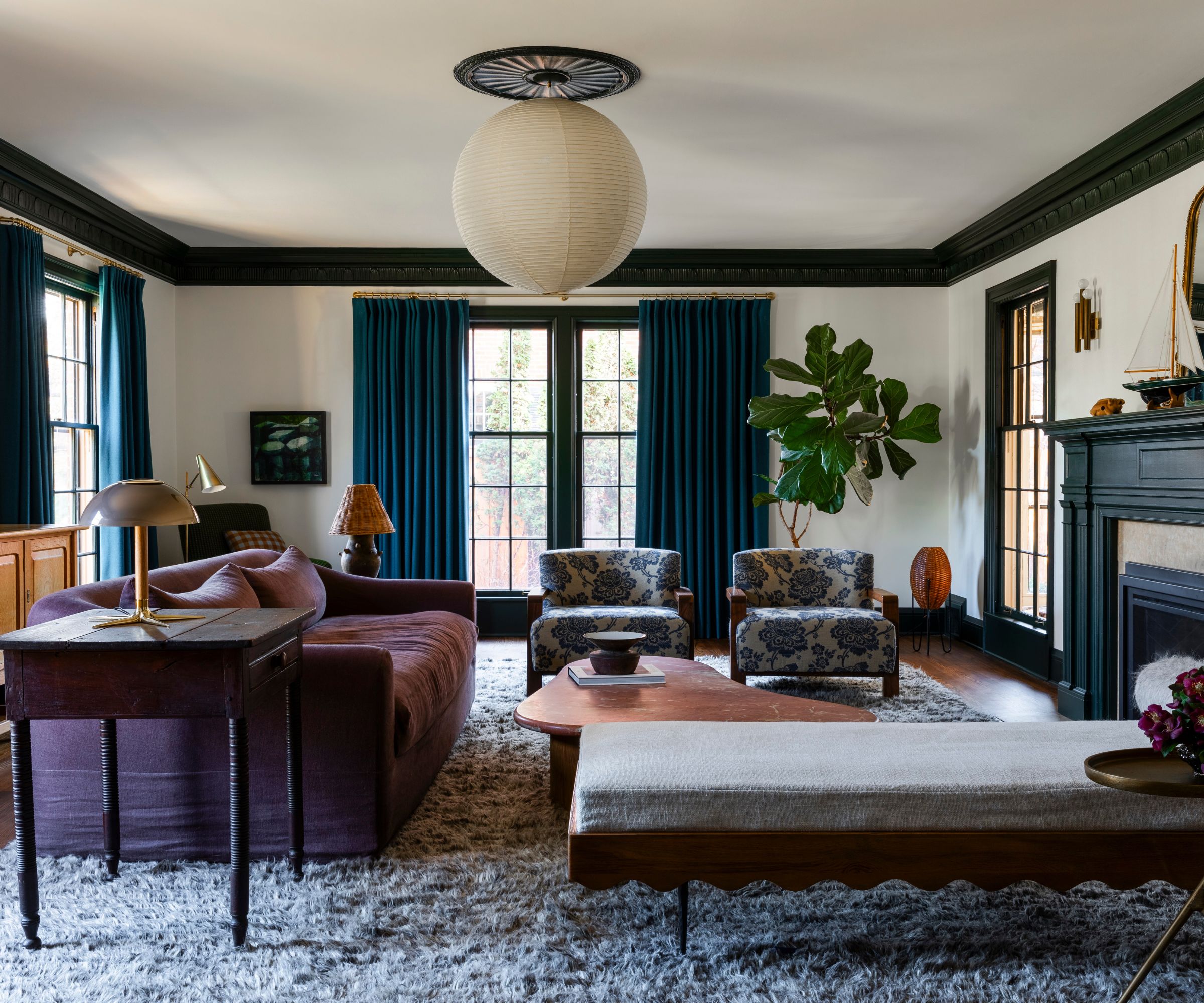 Living room with green ceiling trim, large windows with blue drapes, a large fireplace with a boat on the mantel, a purple sofa, a chaise lounge, and two floral armchairs, with a lantern light above.