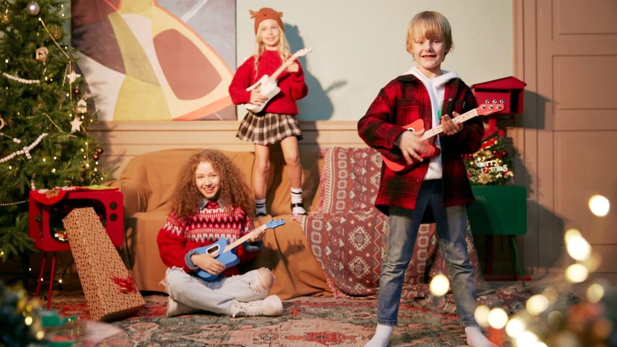 Two girls strumming Tri-Pop guitars, one sitting on couch with red guitar. Older girl on floor with blue guitar. really easy-to-learn