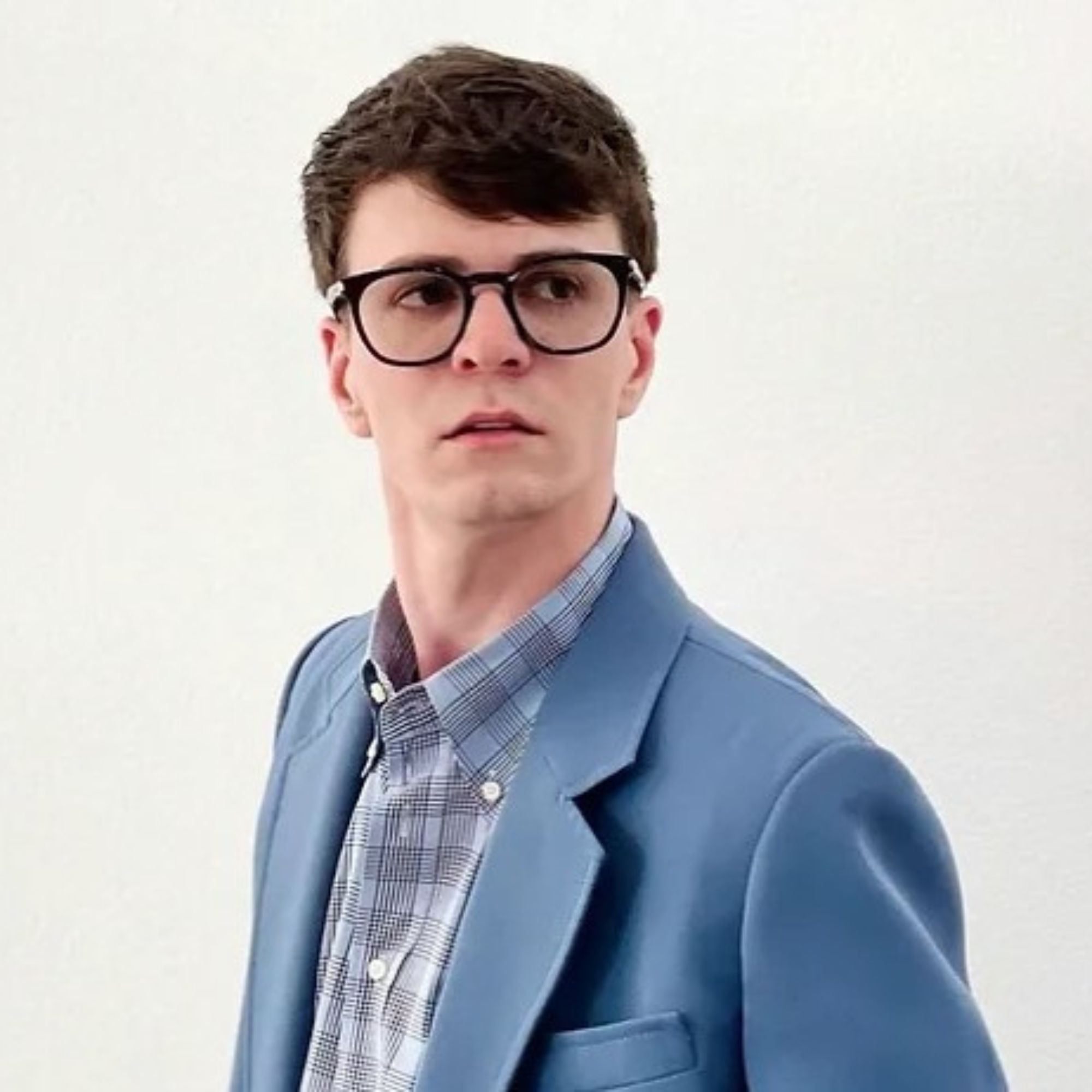 A picture of Michael-Chase Strollo, a man wearing a blue checked shirt and blazer with brown hair and black glasses
