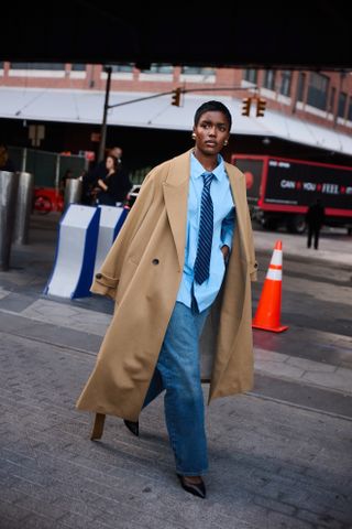 A woman wearing a fall outfit idea of blue jeans, a blue button-down shirt, striped navy tie, and trench coat walking on the sidewalk at Paris Fashion Week