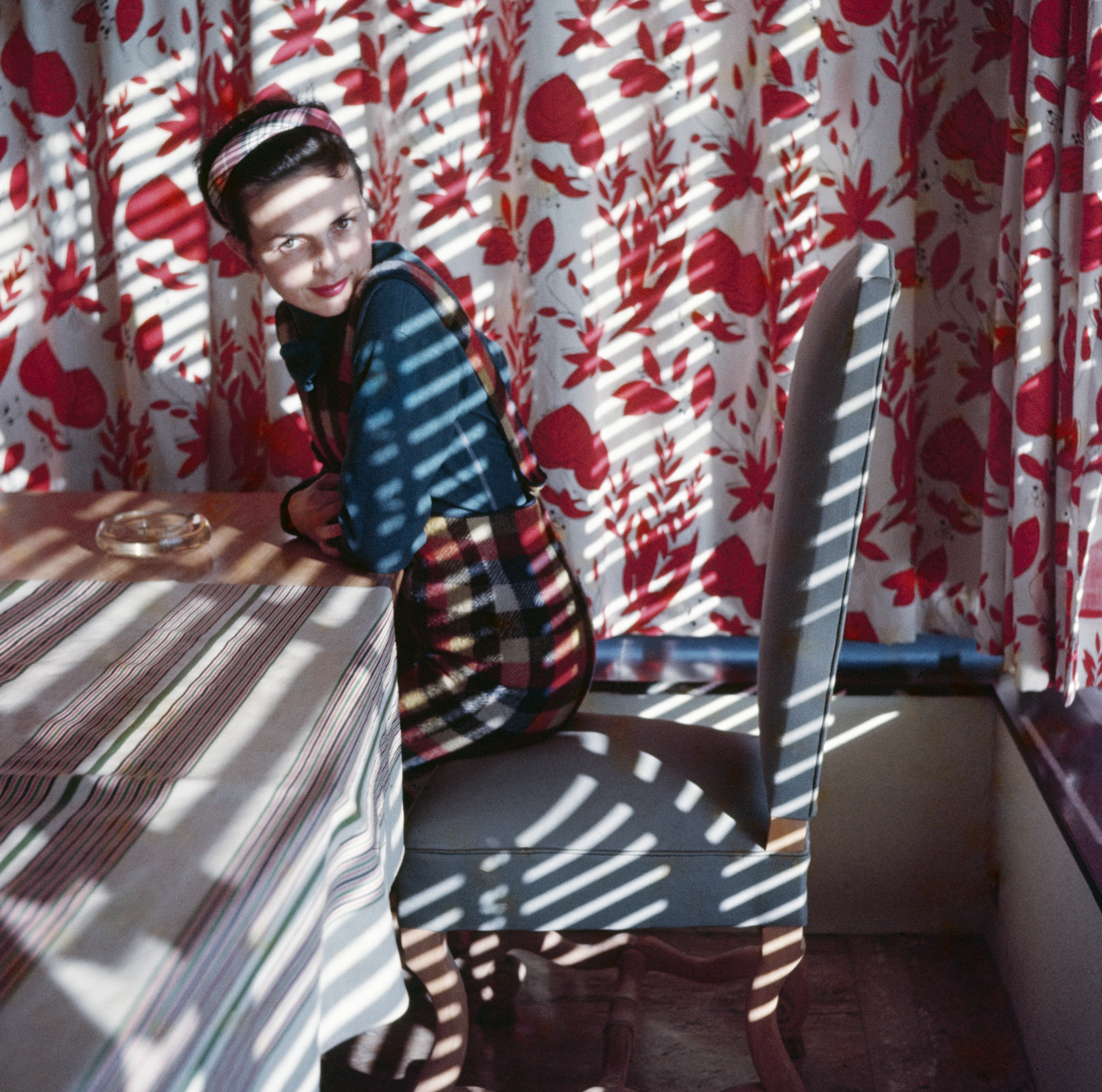 A woman in a tartan dress leans on a striped tablecloth, looking over her shoulder, as window-blind shadows fall across a room hung with red floral curtains.