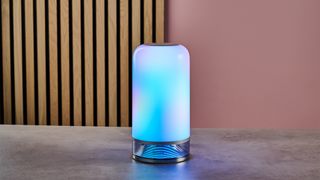 The Govee Table Lamp 2 is lit up with blue and purple colors and sits on a stone surface with wooden slats and a pink wall behind.