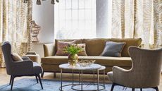 what color curtains are best for summer, living room with caramel/sand leafy patterned drapes, caramel colored couch, marble and metal round coffee table nest, blue rug, oatmeal armchair, colored cushions, vase of flowers 