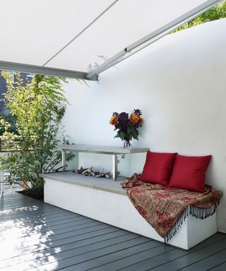 Shaded roof garden ideas featuring white built-in bench seating with red cushions on the side of a gray decked rooftop terrace.