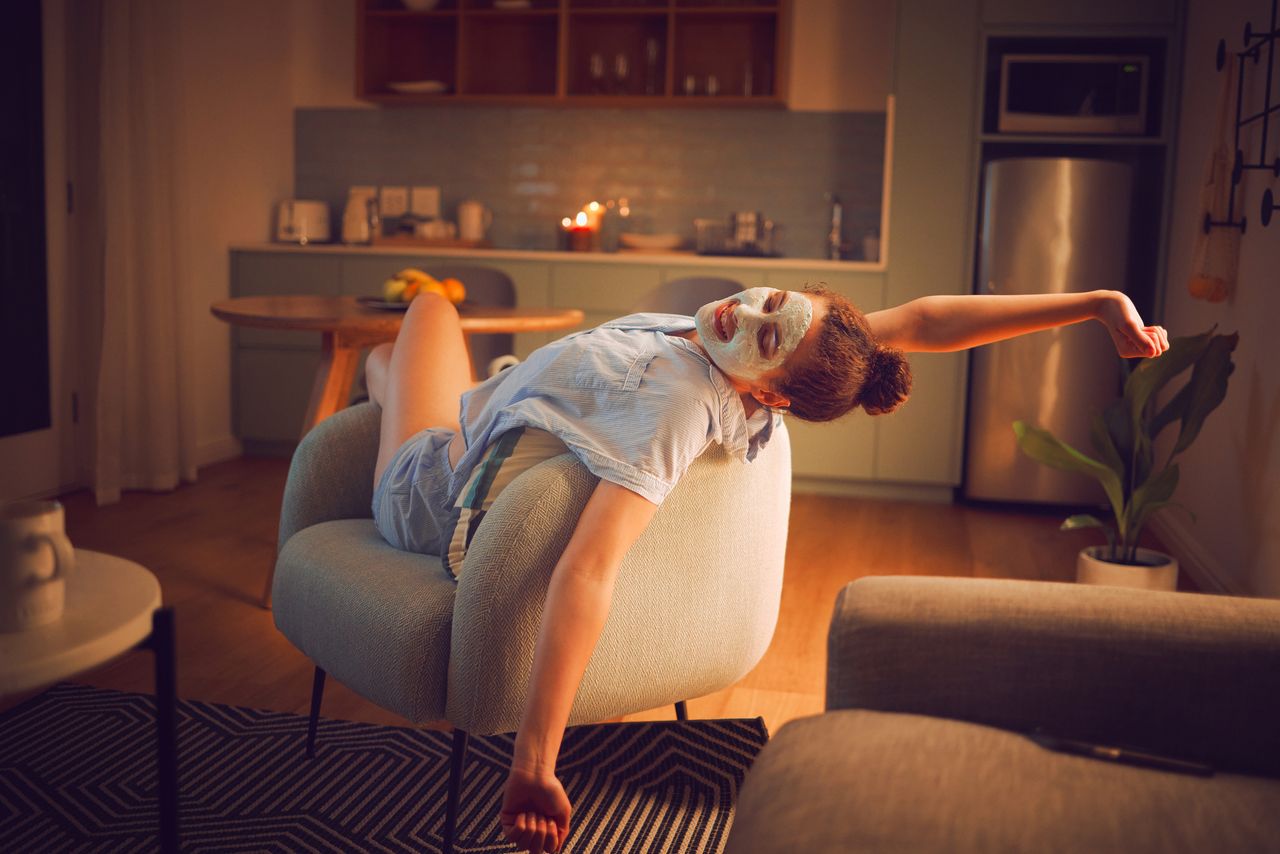 A young woman relaxing in her home with a face mask on and a pair of blue pyjamas, along with low light and lit candles.