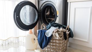 An image of washing machine with an open door and a basket of washing in front of it with a bottle of detergent