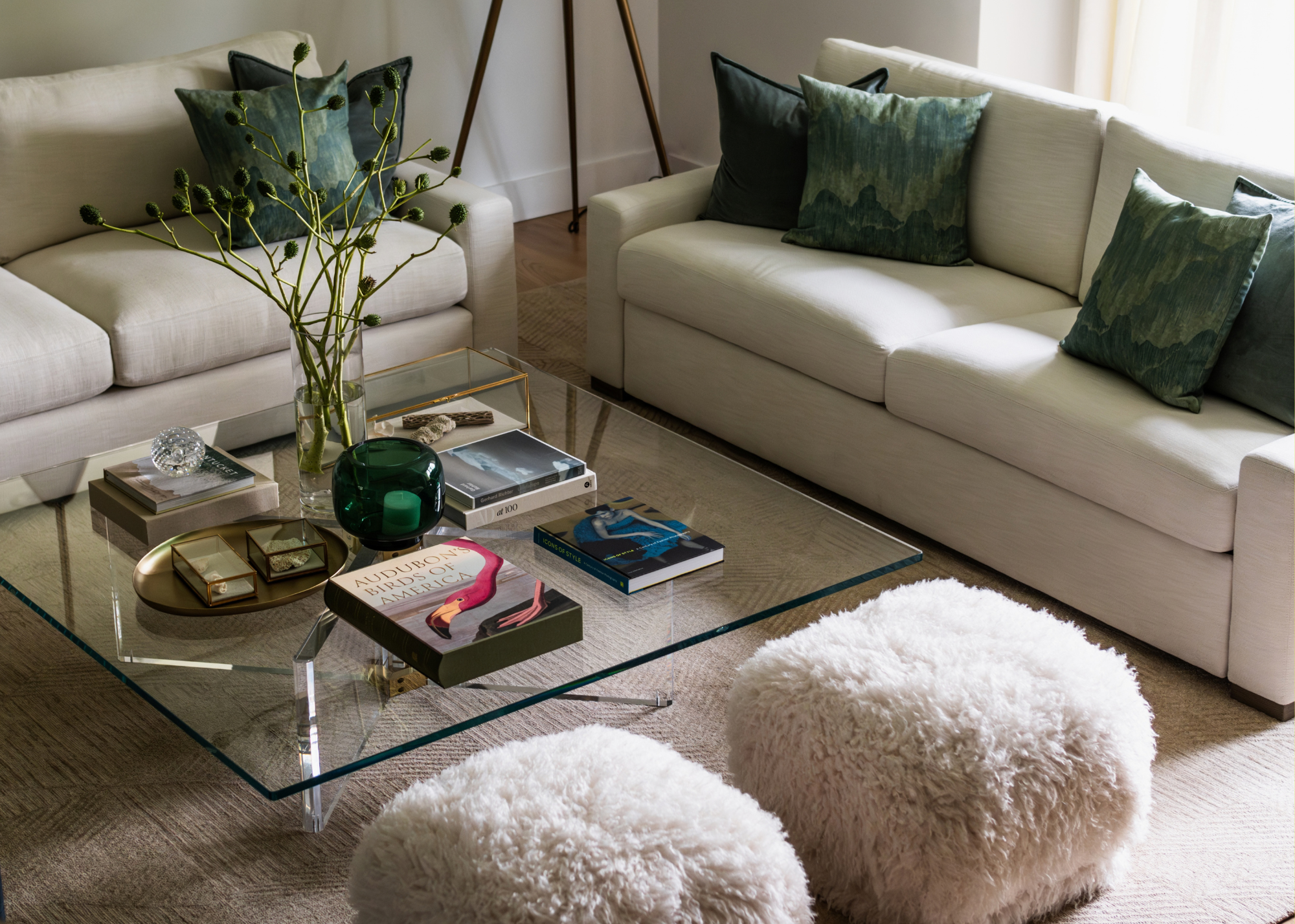A living room with an off-white pair of sofas, a set of fuzzy stools, and a glass coffee table