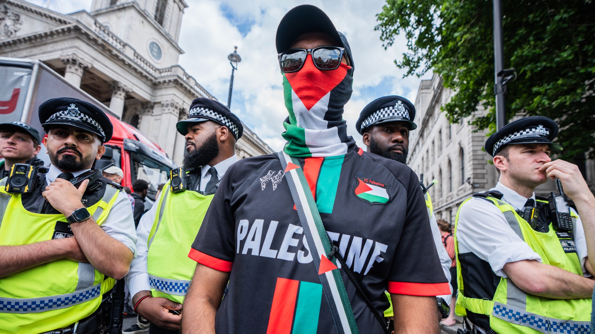 A protestor wearing a Palestinian flag neckscarf and shirt flanked by police officers at a demonstration in Trafalgar Square