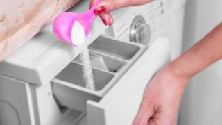 A scoop filled with powder laundry detergent being tipped into a washing machine dispenser drawer