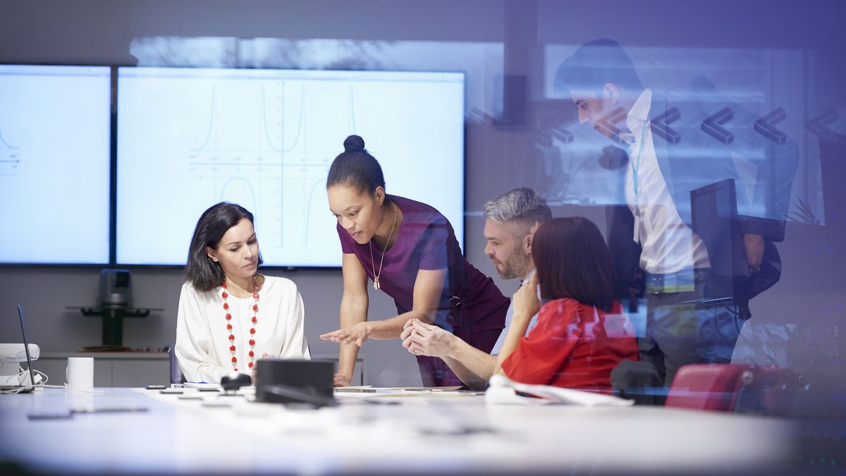 Five business people gathered round a table looking at some plans that are laid flat on the table&#039;s surface. In the background there are some graphs.