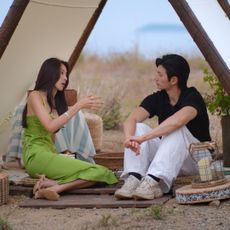 A woman in a green dress (Mina Sue Choi) and a man in a black shirt and white pants (Samuel Lee a.k.a. Lee Sung-hun) talk while sitting under a triangular tarp with wooden posts, decorated with rattan mats, candles in wire cages, and a blue-and-white blanket. A still from 'Single's Inferno' season 5.