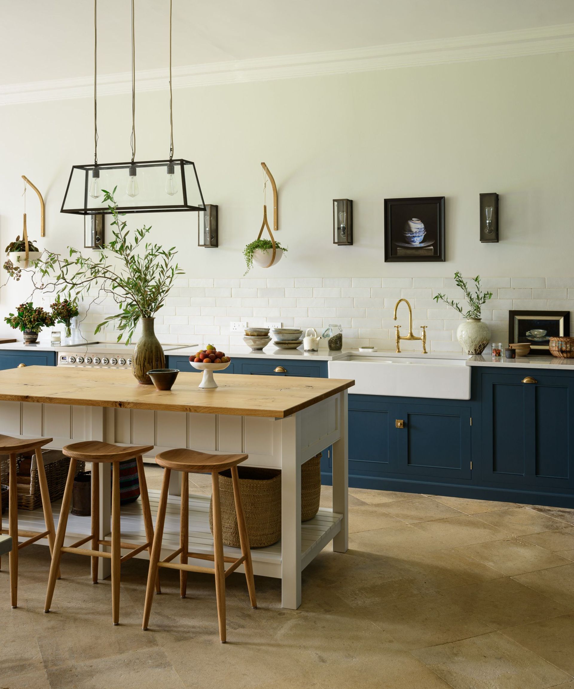 An open plan kitchen with blue lower cabinets, white painted brink walls, and a cream and wood work table as an island
