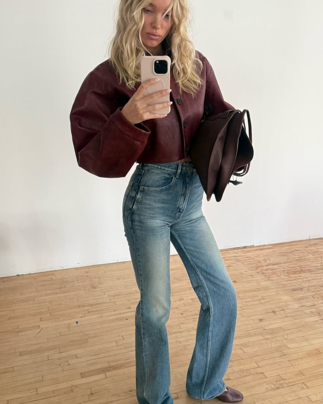 Elsa Hosk takes a mirror selfie wearing blue jeans with a burgundy leather jacket and burgundy flats.