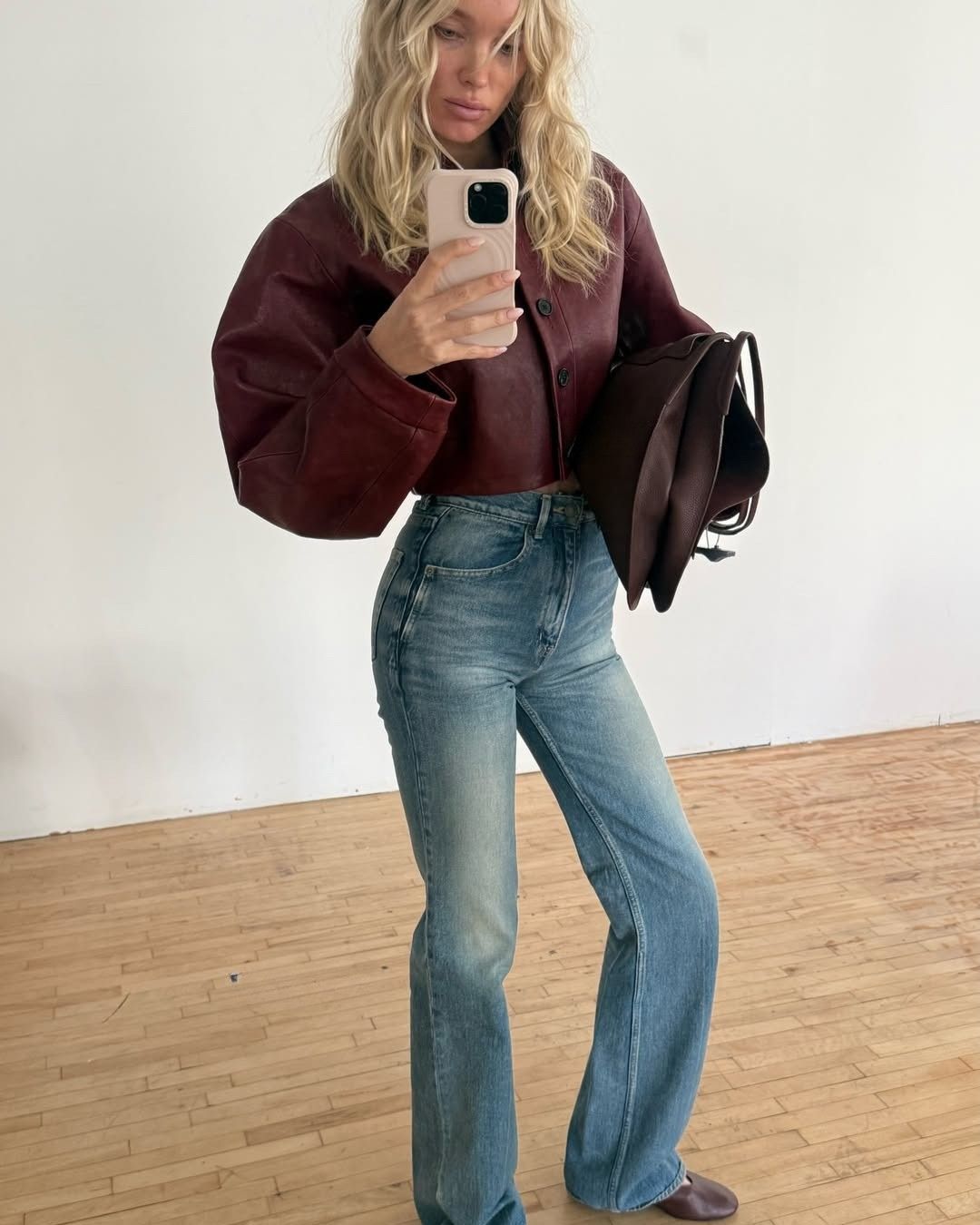 Elsa Hosk takes a mirror selfie wearing blue jeans with a burgundy leather jacket and burgundy flats.