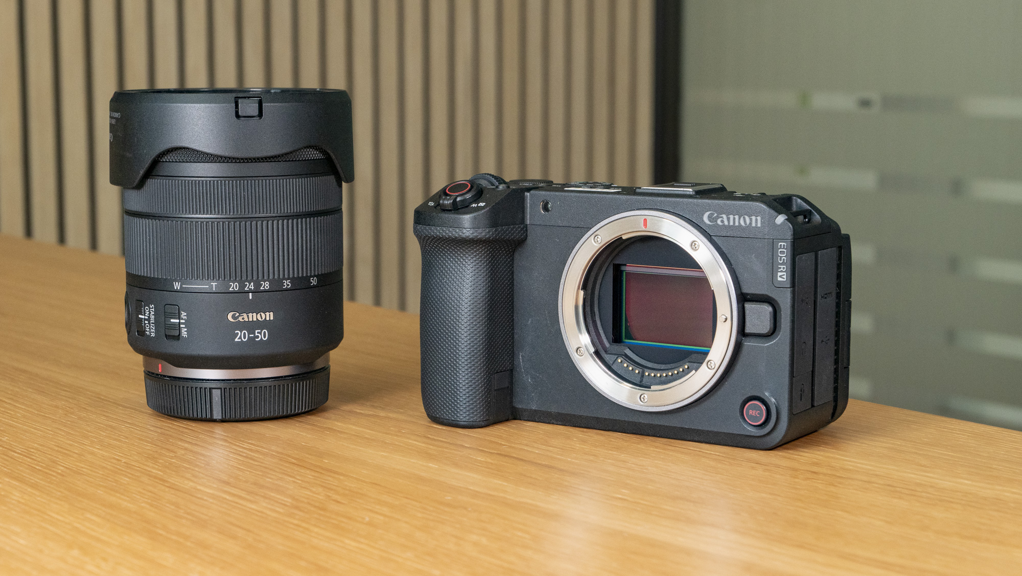 The Canon EOS R6 V mirrorless camera on a wooden table with a pine-slatted backdrop, no lens attached, alongside the RF 20-50mm F/4 L PZ lens
