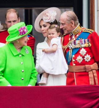 Queen Elizabeth wears a neon green suit and matching hat, while she smiles at a little Princess Charlotte, wearing a white dress, a pink hair clip, and pink shoes