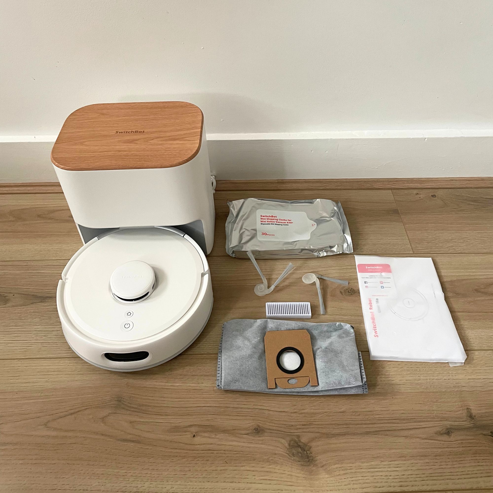 SwitchBot K11+ Robot Vacuum Cleaner with Mop sitting on wood-effect flooring, with accessories laid out next to it