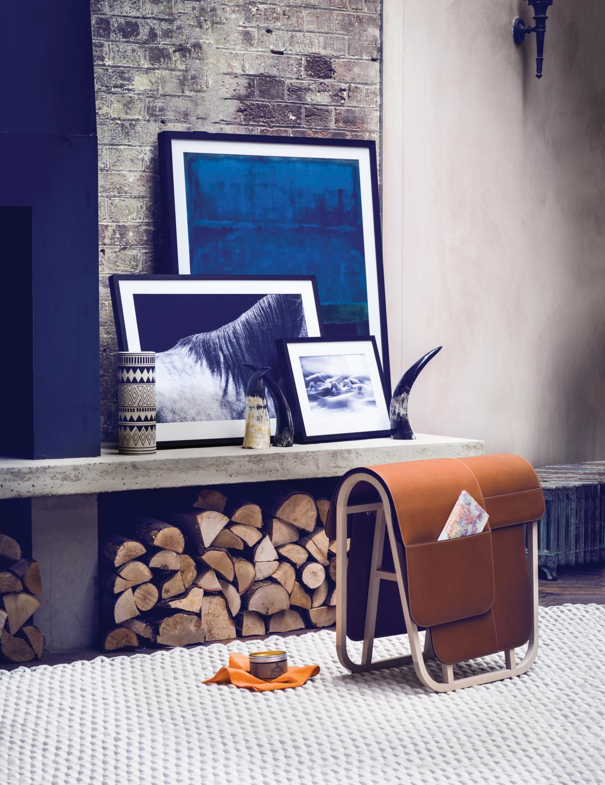 Image of a fireplace area with stacks of chopped wood, a concrete built-in bench next to the fireplace that has paintings of horses leaning on it, and a leather magazine rack.