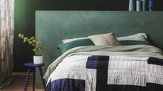 Image of a bed with green, blue, and white bedding. There is a sage green, extended headboard behind it and the walls are a darker shade of green.