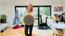 Personal trainer Maddy Biddulph smiles and holds up a wobble board in her living room. Behind her is a Christmas tree, an exercise bike and large windows. 