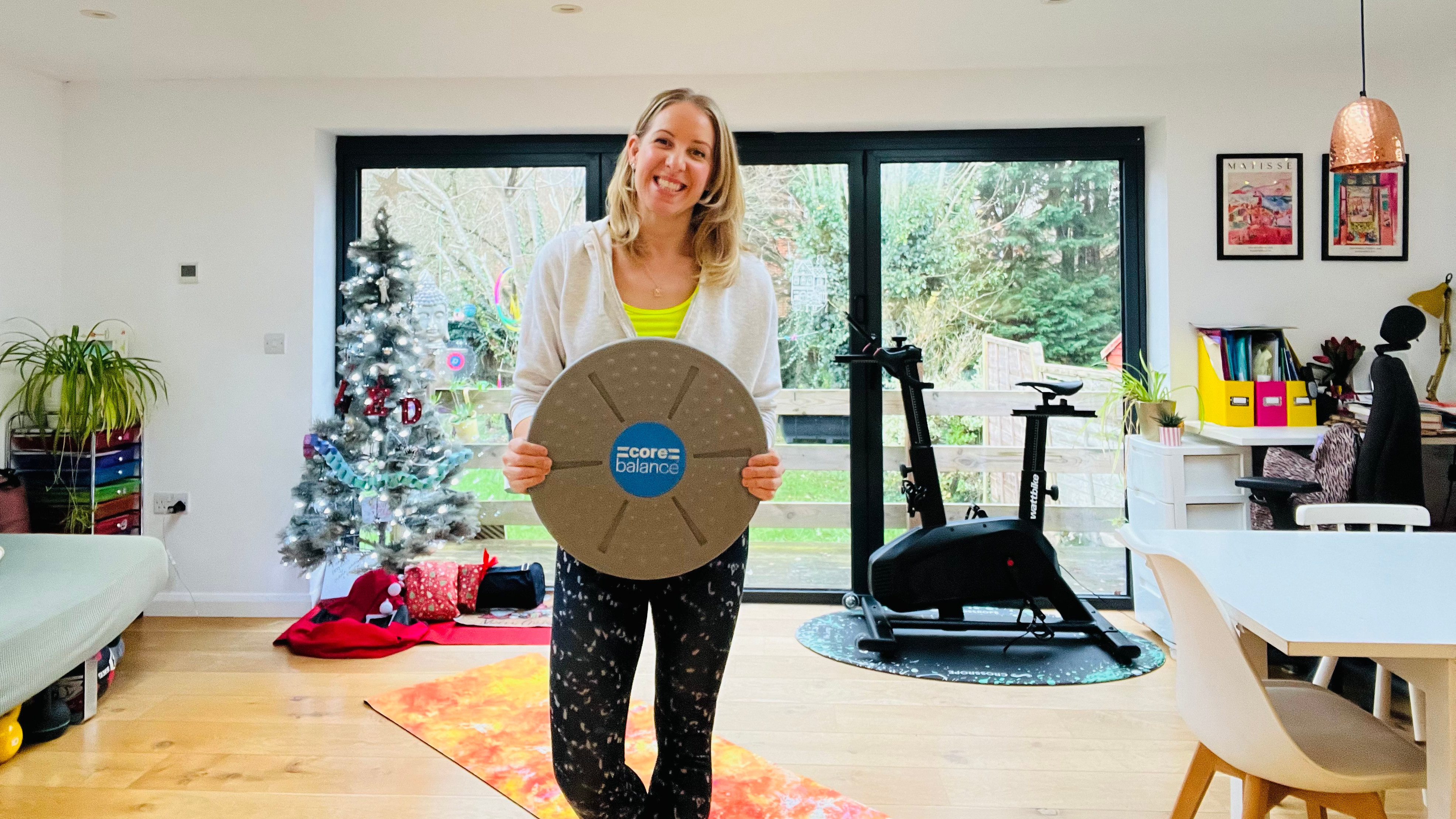 Personal trainer Maddy Biddulph smiles and holds up a wobble board in her living room. Behind her is a Christmas tree, an exercise bike and large windows. 