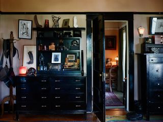 Interior room, two pieces of black furniture with drawers, ornaments and pictures on display, wooden floor witha colourful rug, black door open looking into next room, white walls, black rail framing the top of the walls, wooden coat rail with leather handbags, wall light lit