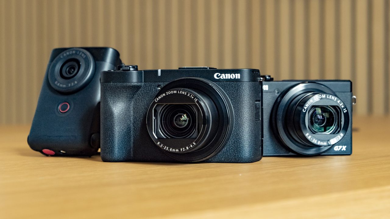 Canon PowerShot V1 compact vlogging camera on a wooden desk alongside the PowerShot G7X Mark III and PowerShot V10