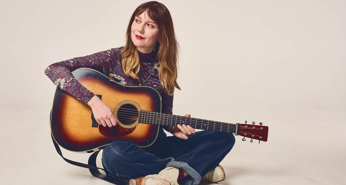 A portrait of Molly Tuttle wearing a patterned shirt, holding a Pre-War dreadnought