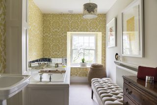 bathroom with yellow damask style wallpaper, built in tub, basket, upholstered footstool, cabinet, mirrored splashback