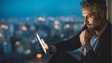 A businessman looks at a tablet against the backdrop of a nighttime skyline.