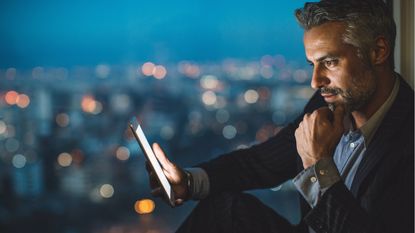 A businessman looks at a tablet against the backdrop of a nighttime skyline.