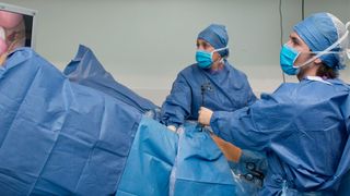 two medical providers dressed in face masks and blue surgical scrubs examine a patient (covered in a blue sheet) during a surgery to evaluate for endometriosis