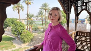 Lucy Worsley in a pink top stands on a balcony overlooking palm trees.