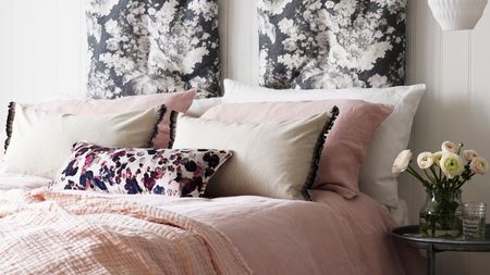 A white panelled bedroom with a double bed with pink bedding and cushions beside a black circular nightstand with a vase of flowers