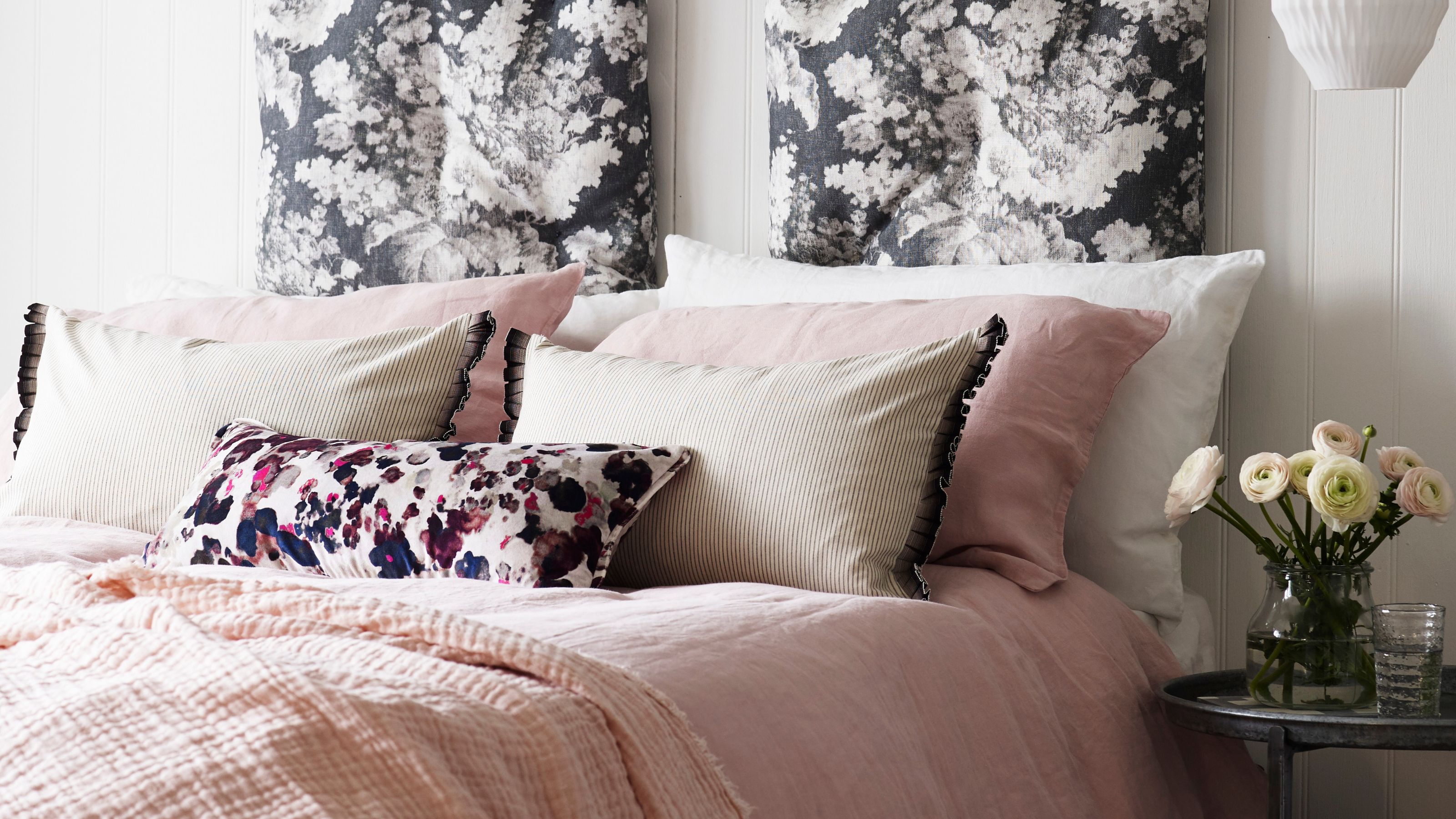 A white panelled bedroom with a double bed with pink bedding and cushions beside a black circular nightstand with a vase of flowers