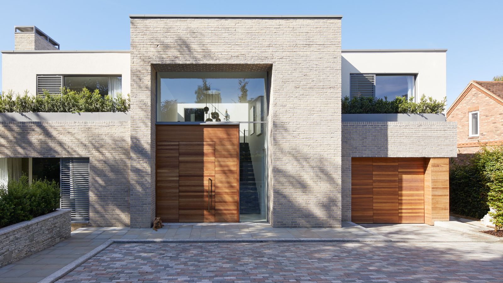 Contemporary home with a pale brick exterior, large wooden front door, floor-to-ceiling glass, and a paved driveway with multicoloured stone bricks.