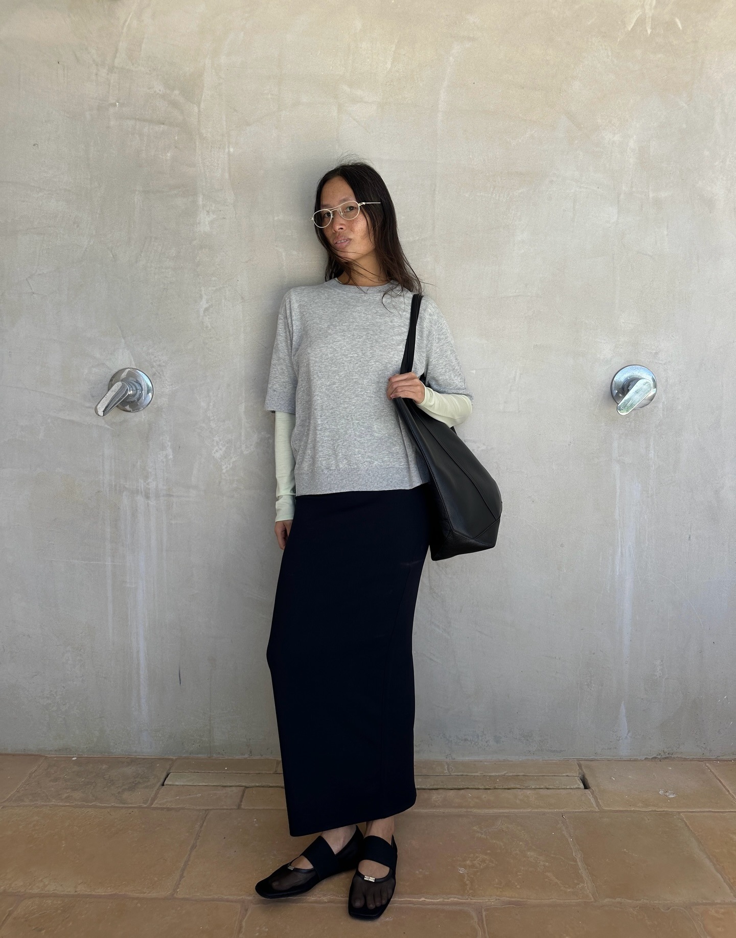 French woman wearing a travel outfit that includes a gray tee layered over a long-sleeve one, a black column skirt, and black ballet flats
