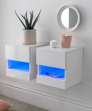 Contemporary bedroom setup with sleek, wall-mounted white bedside units featuring illuminated blue shelving. The minimalistic design is complemented by a potted aloe plant and candles