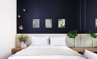 A room in the MyChelsea hotel. Deep blue and white walls, with plant art in golden frames hanging above the bed. The bed is covered in white linen, with a beige headboard, and a wooden board that serves as a night table, that has plants on it. The mirror covers the right wall.
