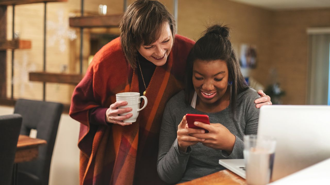 A mother and daughter look at the daughter&#039;s phone together while the daughter sits in front of a laptop on the dining room table.