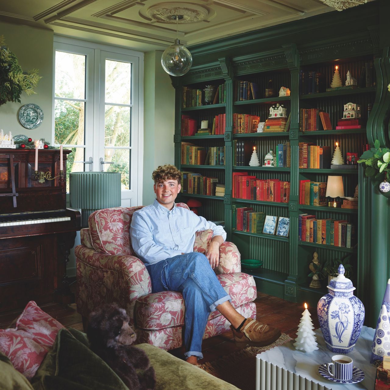 content creator Jack Kinsey sitting in dark green library room with bookshelves around fireplace with woodburner and fluted marble-look coffee table