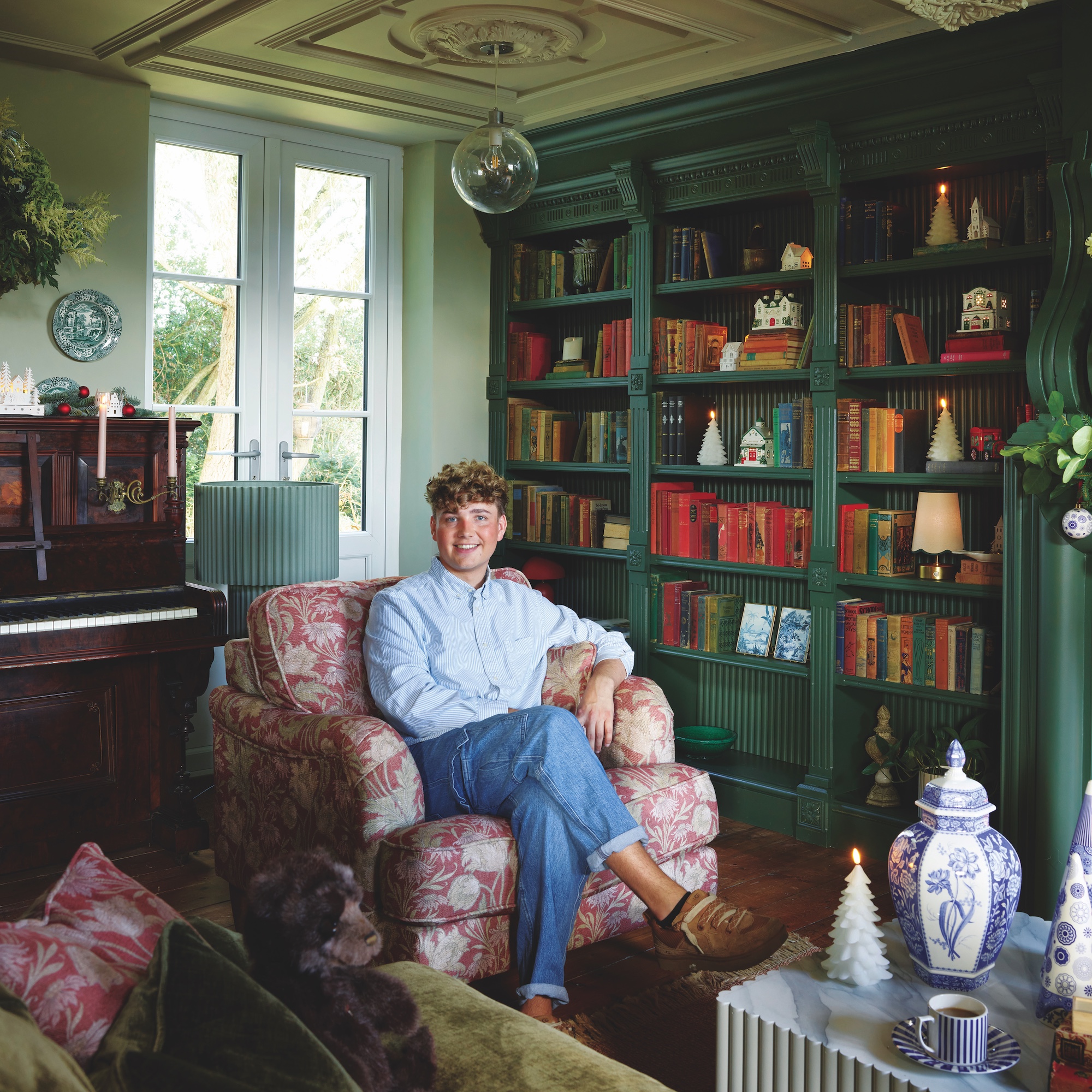 content creator Jack Kinsey sitting in dark green library room with bookshelves around fireplace with woodburner and fluted marble-look coffee table