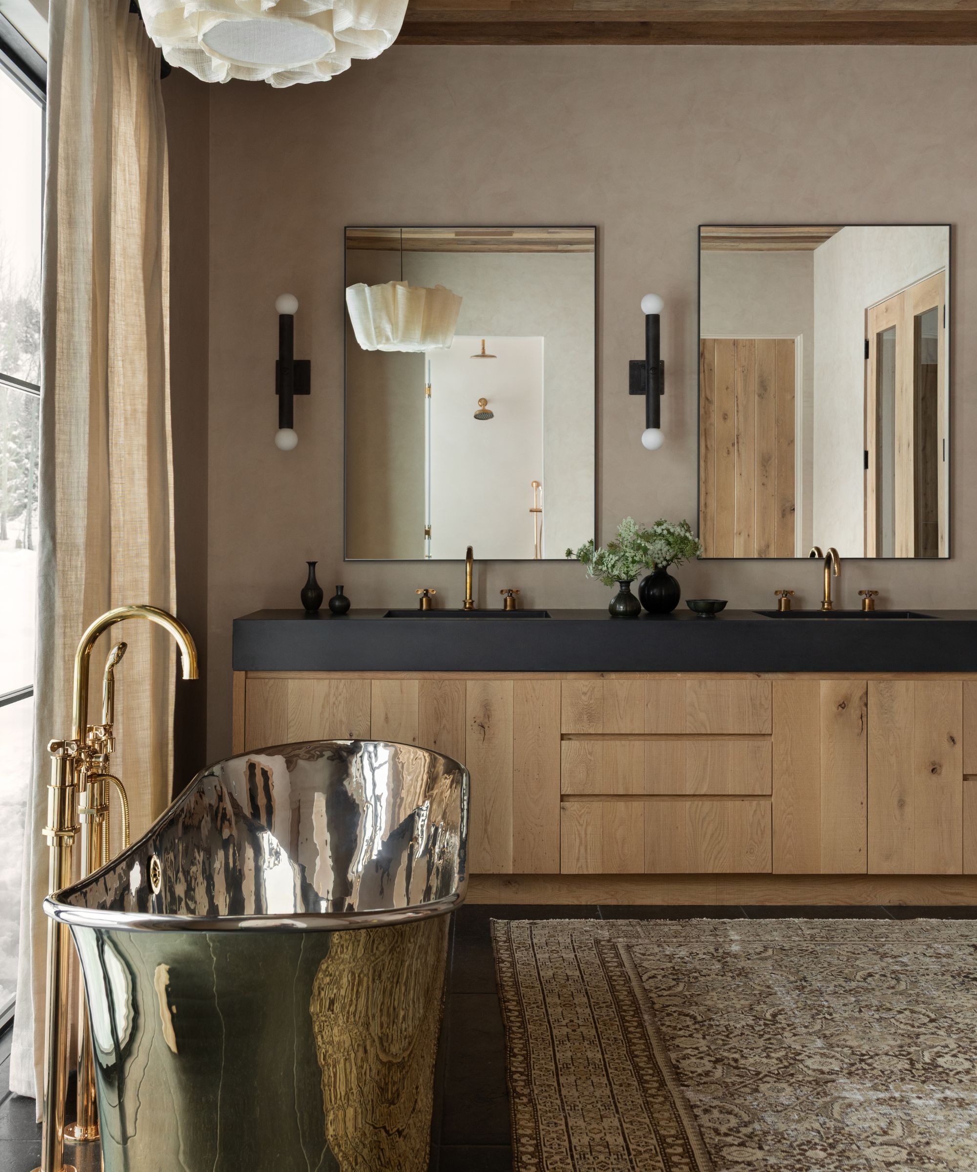 a neutral bathroom suite with oak double vanity, dark counters, two mirrors, a vintage rug and a freestanding brass tub