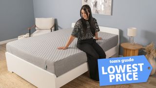 A woman sits on the edge of the Siena Memory Foam Mattress in a bedroom, testing the edge support of this cheap gel memory foam mattress. A Tom's Guide lowest price deals graphic in the right corner