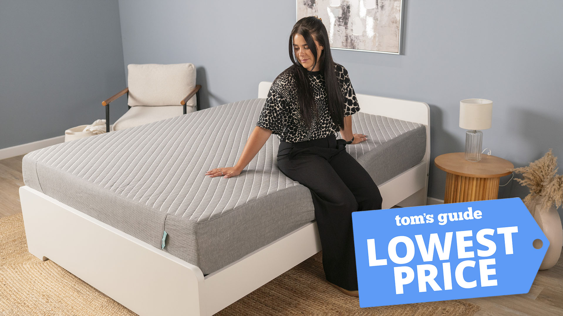 A woman sits on the edge of the Siena Memory Foam Mattress in a bedroom, testing the edge support of this cheap gel memory foam mattress. A Tom&#039;s Guide lowest price deals graphic in the right corner
