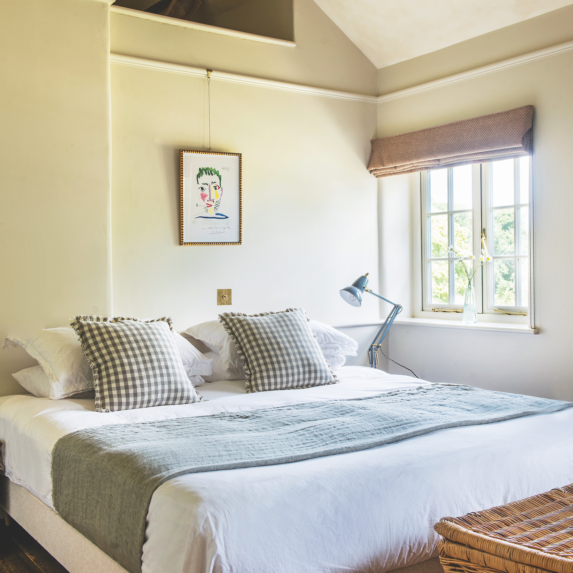 simple bedroom with cream walls, bed with check cushions and window with roman blind